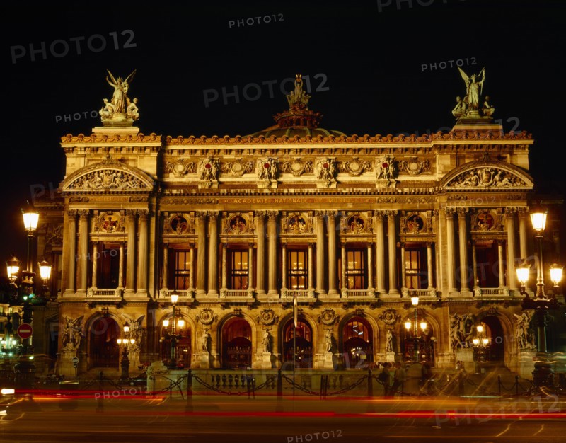 FRANCE, Ile de France, Paris, Opera frontage floodlit at night against black sky with light trails from passing traffic in the foreground.
