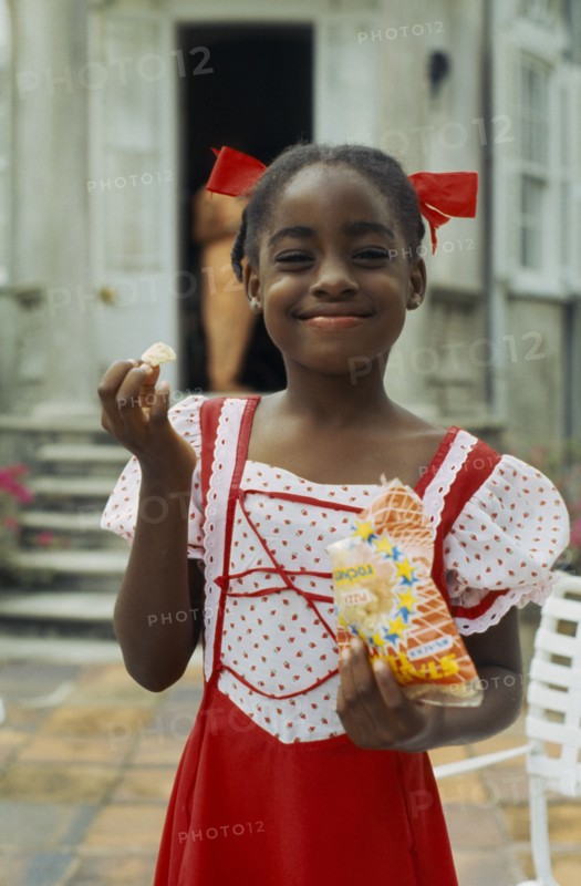 WEST INDIES, Barbados, Easy Hall, Young smiling girl in red and white dress with red ribbons in hair eating crisps from a packet