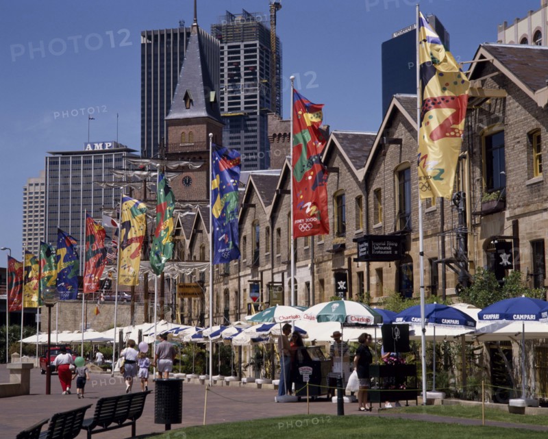 AUSTRALIA, New South Wales, Sydney, The Rocks harbourside area with people walking past restaurants festooned with umbrellas and flags