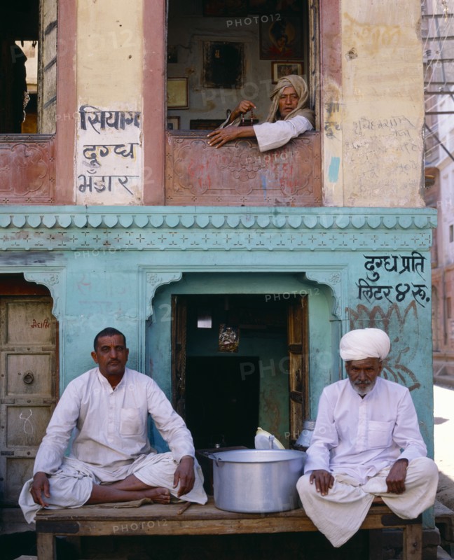 INDIA, Rajasthan, Bikaner, Two men sitting on table outside shop with man framed  in window above.