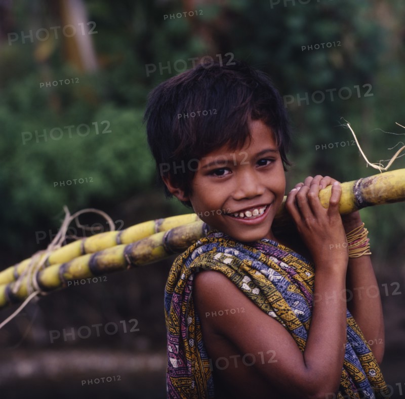 INDONESIA, Lombok, Kuta, Smiling boy with a sarong over his shoulder carrying sugarcane