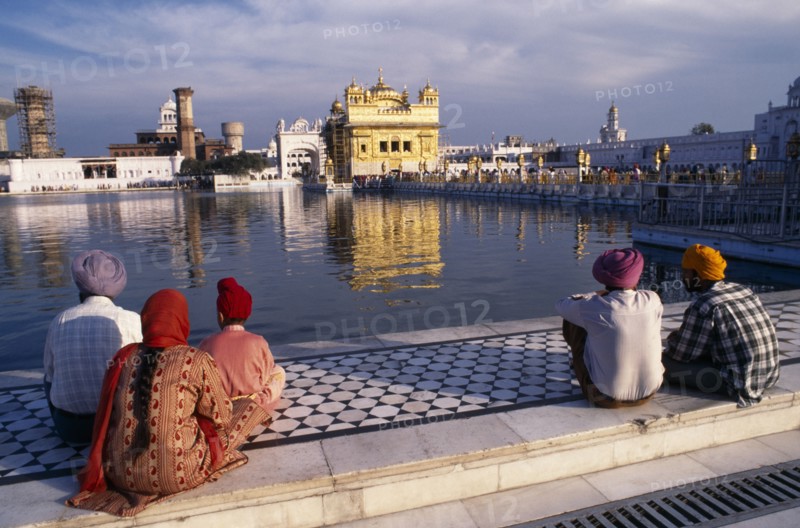 INDIA, Punjab, Amritsar, "Sikh men, woman and child sitting on marble walkway beside the sacred pool looking towards the Golden Temple reflected in the water."