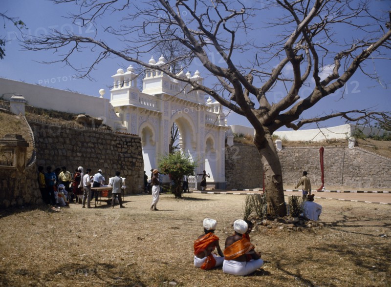 INDIA, Karnataka, Mysore, "Maharaja’s Palace.  Exterior wall and white, carved and decorated entrance arch with people gathered in dry, grassy area in foreground."
