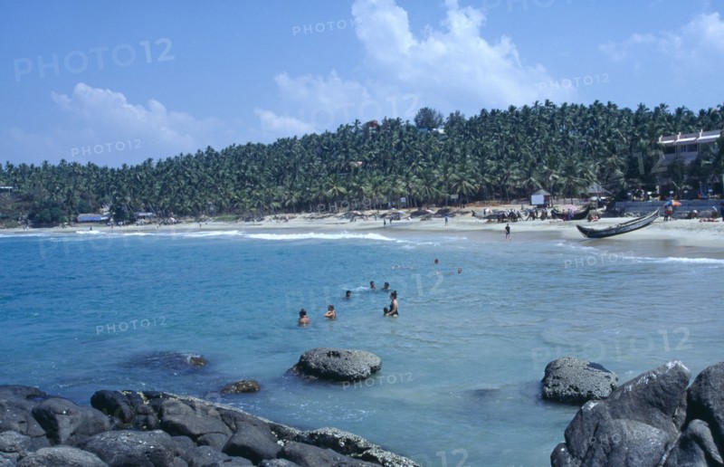 INDIA, Kerala, Kovalam, View from rocky outcrop over bay toward beach surrounded by palms with group of bathers in the sea
