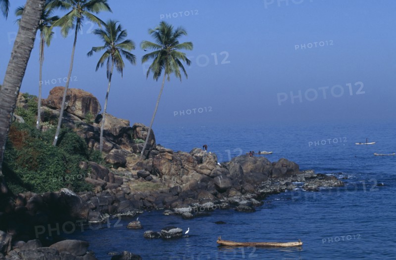 INDIA, Kerala, Kovalam, Coastal scene with coconut palms on rocky promontory and men in boats beyond