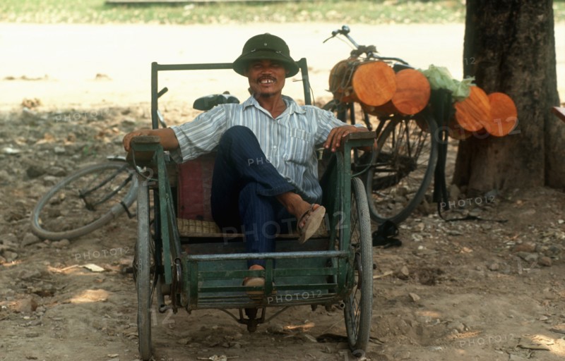 VIETNAM, Hanoi, Cyclo driver sitting in his vehicle under the shade of a tree.