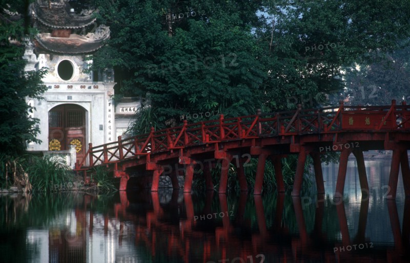 VIETNAM, Hanoi, Hoan Kiem Lake. View of red painted bridge across water to decorative building surrounded by lush vegetation.