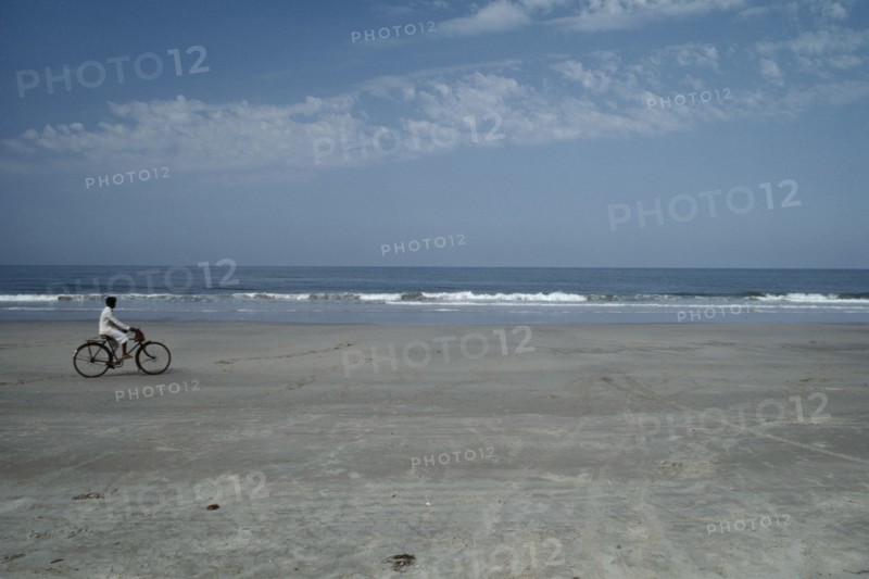 INDIA, Goa, Colva Beach , Cyclist riding along sand of the empty beach with waves rolling on to the shore