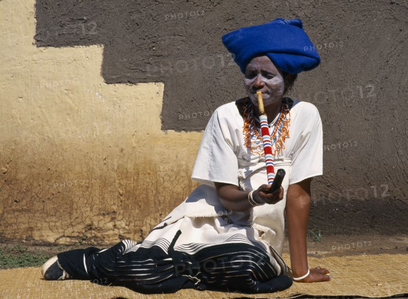 SOUTH AFRICA, Lesedi, Xhosa lady smoking pipe on street with head wear and painted face.