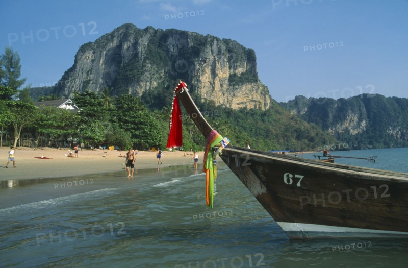 THAILAND, Krabi, Ao Nang Beach, Long tail boat moored in shallow water at the edge of beach with tourists.  Forest covered cliffs behind.