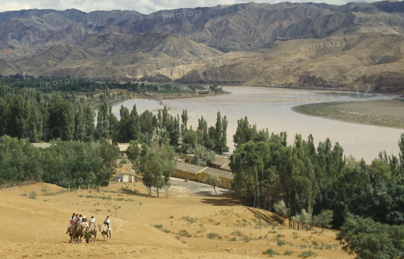 CHINA, Ningxia , Yellow River, "View over river bend in sandy region of north west China.  Group of people on camels in the foreground, mountains behind."