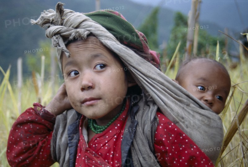 NEPAL, Bung, Rai girl carrying baby in sling on her back supported by cloth knotted around head.