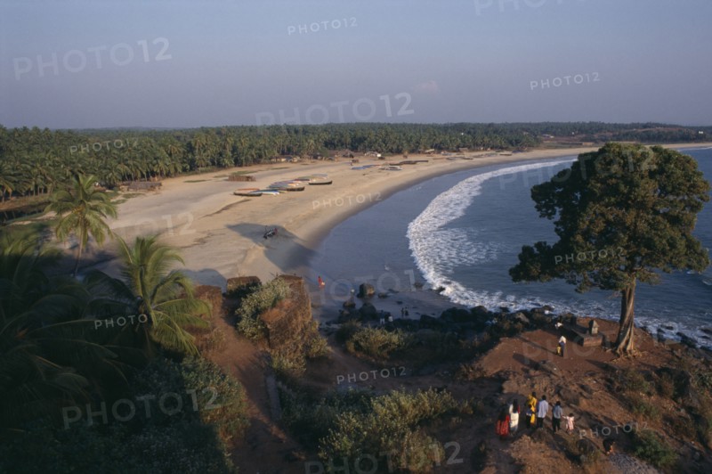INDIA, Kerala, Bekal, View along the beach and coastline with people standing by tree on rocky promontory in the foreground