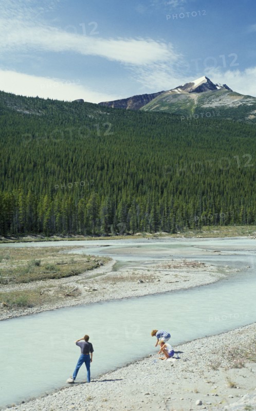 CANADA, British Columbia, Jasper National Park, Visitors beside river looking towards dense pine forest with jagged mountain peak above.