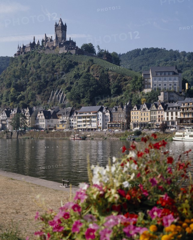 GERMANY, Rhineland, Cochem, Hilltop castle overlooking houses beside the Mosel River with flowers in the foreground