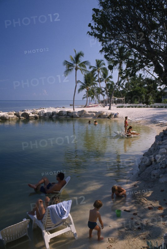 USA, Florida, Islamorada, "Cheech Lodge Beach with palms, people sitting on sun loungers in the water and children playing in the foreground"