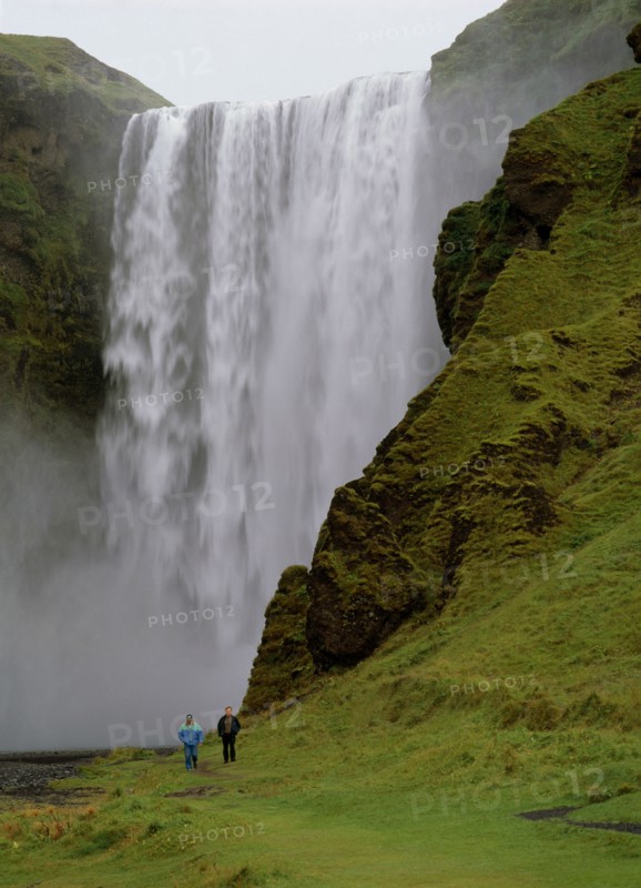 ICELAND, Skogafoss Waterfall, Cascading waterfall 60 metres or 200 feet high.  Two people walking along footpath at side.