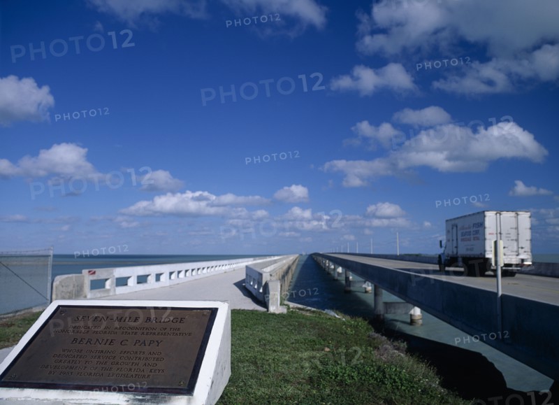 USA, Florida Keys, Seven Mile Bridge. Commemorative plaque with the Honorable Florida State Representative Bernie C Papy seen in the foreground with a truck driving along the concrete bridge stretching out over the sea