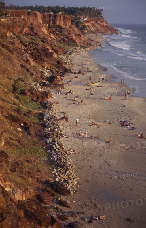 INDIA, Kerala, Varkala, "Beach sheltered by steep, eroded cliffs with sunbathers on the sand and palm trees on headland beyond."