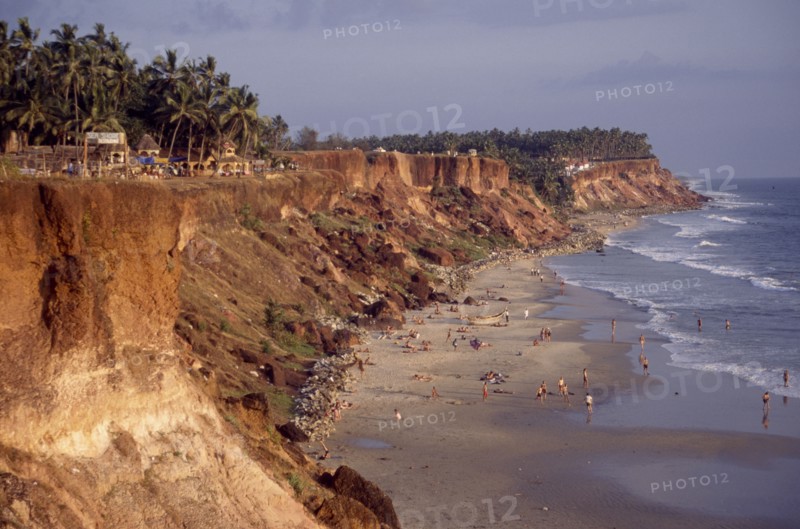 INDIA, Kerala, Varkala , "Coastline and narrow sandy cove with sunbathers sheltered by steep, eroded cliffs with bars and restaurant backed by palm trees along top."