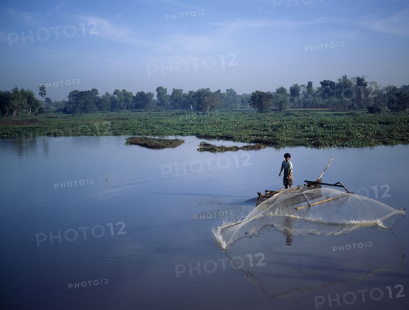 INDONESIA, Lombok, Timur NTB, "Praya, man fishing from outrigger canoe using circular casting net, lake weeds & trees behind J6599 "