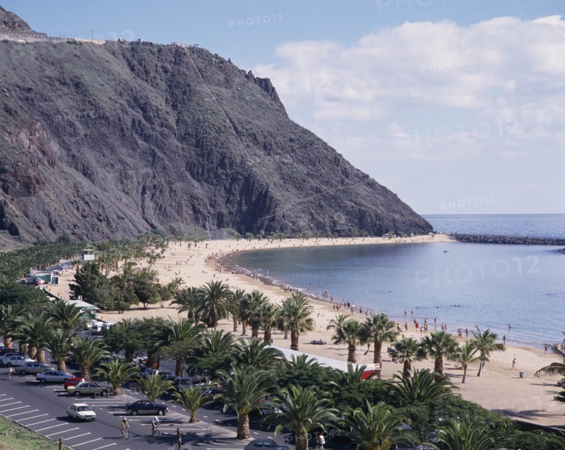 SPAIN, Canary Islands, Tenerife, Las Teresitas. Long sandy bay beneath black cliffs and palm lined promenade with parked cars