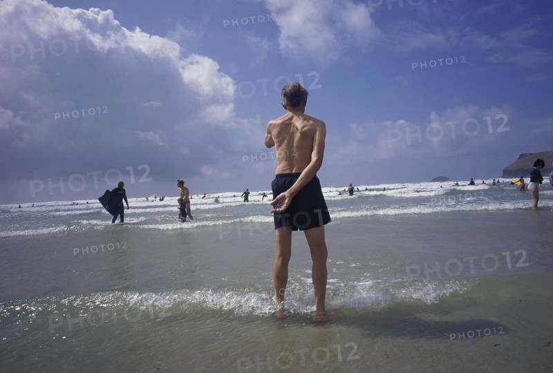 ENGLAND, Cornwall, Polzeath, Man standing in shallow water on the beach looking out to bathers in the sea
