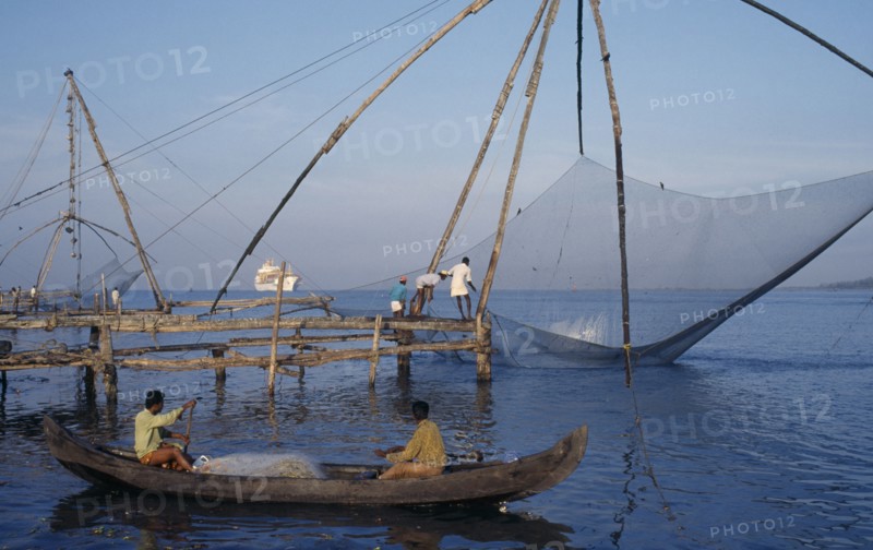 INDIA, Kerala, Kochi, Chinese fishing net being raised with a catch as two men row past in a canoe and a white ship sits on the horizon
