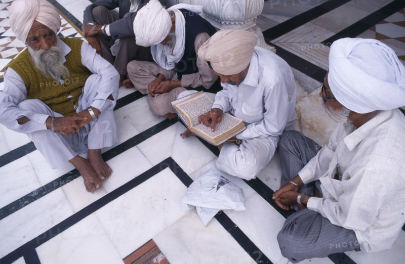 INDIA, Punjab, Amritsar, Group of Sikh men sitting on black and white marble floor of Golden Temple complex to read holy book.
