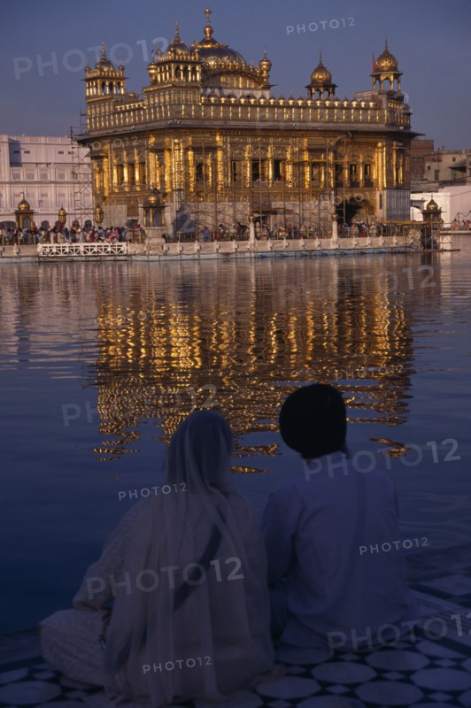 INDIA, Punjab, Amritsar, Sikh couple sitting on marble walkway looking out across sacred pool towards the Golden Temple and shimmering reflection on rippled water surface.
