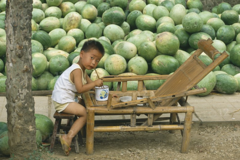 CHINA, Guizhou Province, Kaili, Liitle boy eating from bowl with chopsticks with large pile of watermelons behind.