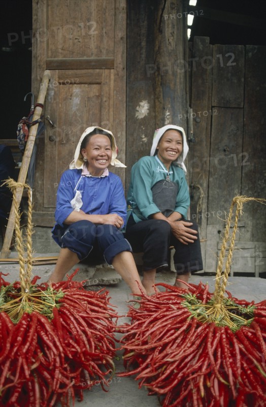 CHINA, Guizhou, Yuno, Two smiling female chilli vendors sitting on a pavement behind their chillies