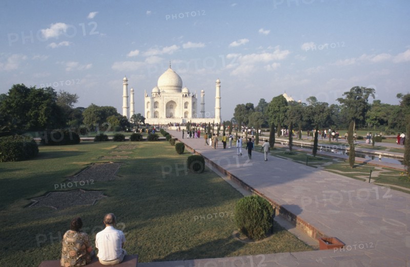 INDIA, Uttar Pradesh, Agra, Taj Mahal and gardens with couple sitting in the foreground and other people on a path leading up to the building
