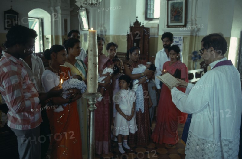 INDIA, Goa, Margao, "Church interior with priest conducting christening service, three women standing holding babies with other guests and family members."