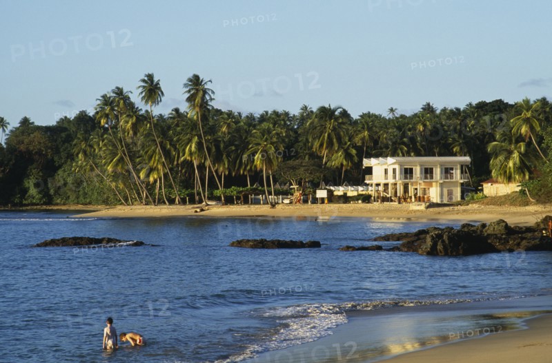 WEST INDIES, Tobago, Grafton Beach, People standing in sea beside sandy beach with white painted building surrounded by palm trees.