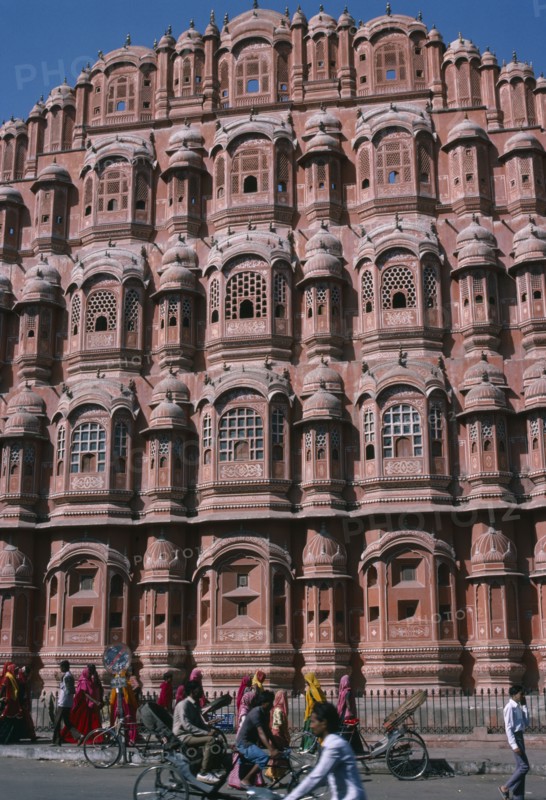 INDIA, Rajasthan, Jaipur, "Hawa Mahal or Palace of the Winds.  Part view of semi-octagonal, honeycombed sandstone facade.   Cyclists and brightly dressed women on street in front. "