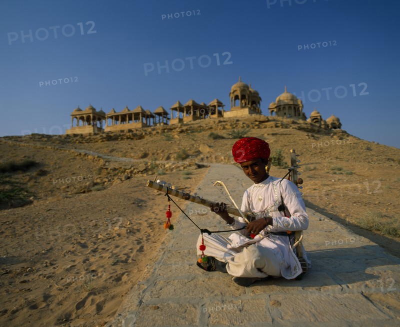 INDIA, Rajasthan, Jaisalmer, Musician wearing a red turban sitting cross-legged on the ground with old buildings on top of the hill behind him