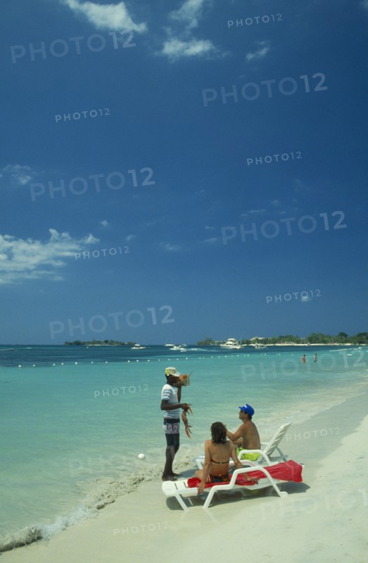 WEST INDIES, Jamaica, Negril, Man selling lobster to tourists on beach sitting on sun loungers by the water