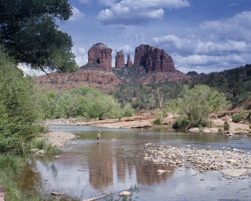 USA, Arizona, Sedona, Cathedral Rock in Oak Creek Canyon. View across river towards wind eroded rock features with a boy wading in the water.