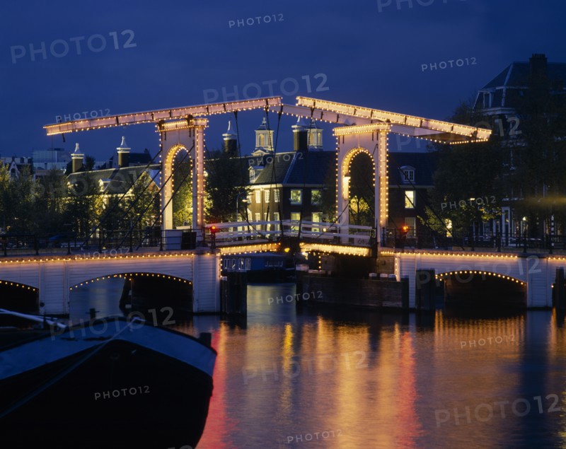 HOLLAND, Noord, Amsterdam, Magere Brug the Skinny Bridge illuminated at night with a blue sky and houses in the  background