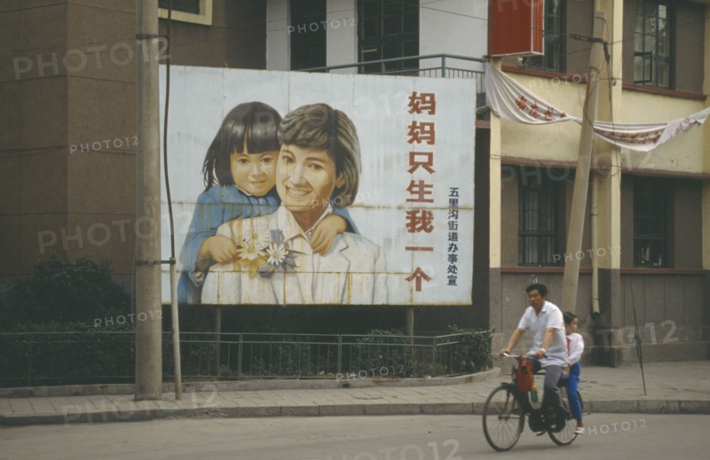 CHINA, Shandong, Jinan, Man with child passenger cycling past birth control poster with image of female child.
