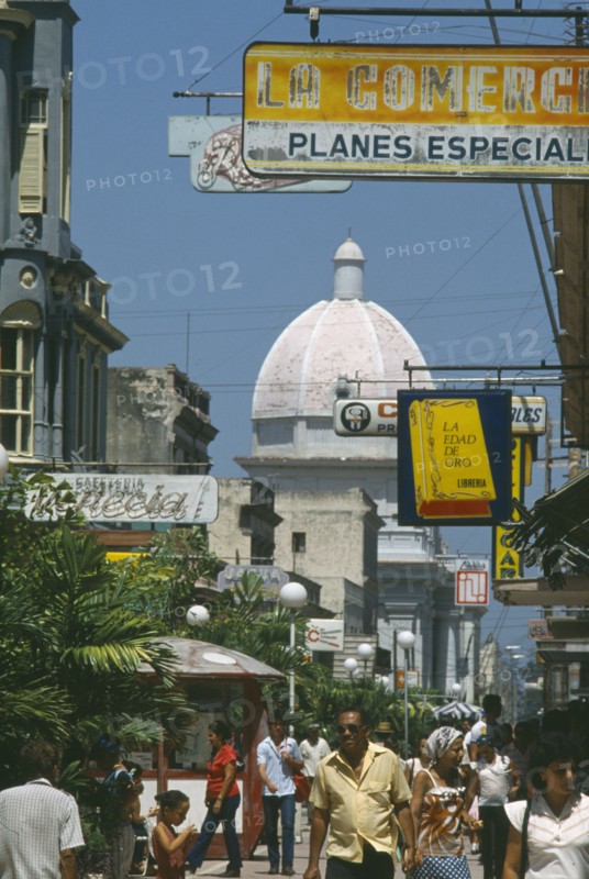CUBA, Cienfuegos, "Busy street scene with people, hanging shop signs and dome of white building beyond."