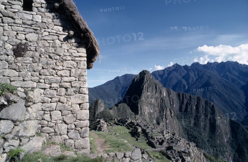 PERU, Cusco, Machu Picchu, View of the side of a stone built thatched house with ruins beyond