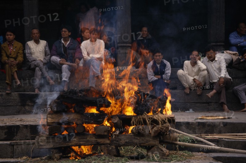 NEPAL, Kathmandu, Hindu cremation in Pashupatinath Temple.  Burning wood funeral pyre with onlookers on steps behind.
