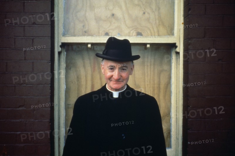 IRELAND,  , Dublin, Priest wearing hat in front of boarded up house window.