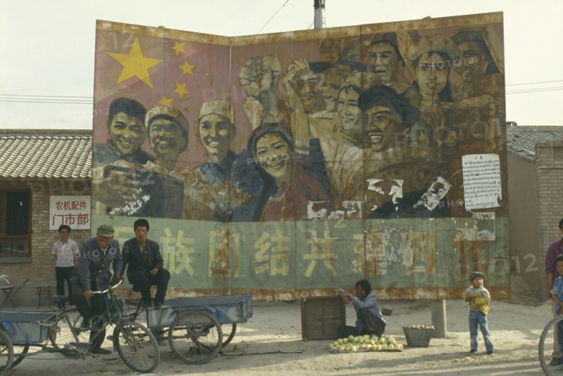 CHINA, Ningxia, Guyuan , Old Communist poster on hoarding at road junction with woman selling fruit below. Child and two men on stationary bicycle driven carts.