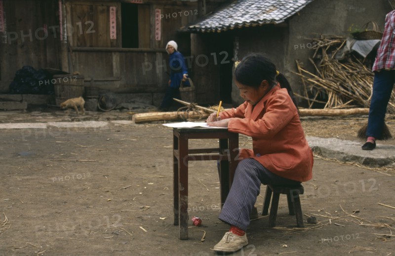 CHINA, Guizhou, Young girl doing homework at desk in farmyard.