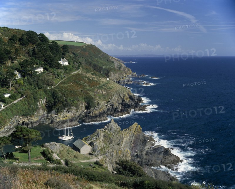 ENGLAND, Cornwall, South Cornwall . Rugged coastline with yachts in bay and houses on hillside
