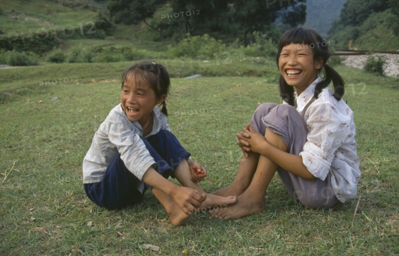 CHINA, Guangxi, Two farm girls sitting on grass laughing.