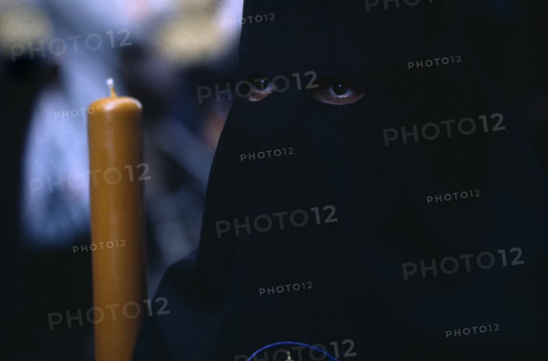 SPAIN, Andalucia, Seville, Semana Santa Easter procession. Penitent in black robe with eyes visable through hood holding a candle